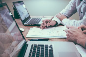 Two people sitting at a desk writing on a stack of papers with laptops sitting in front of them